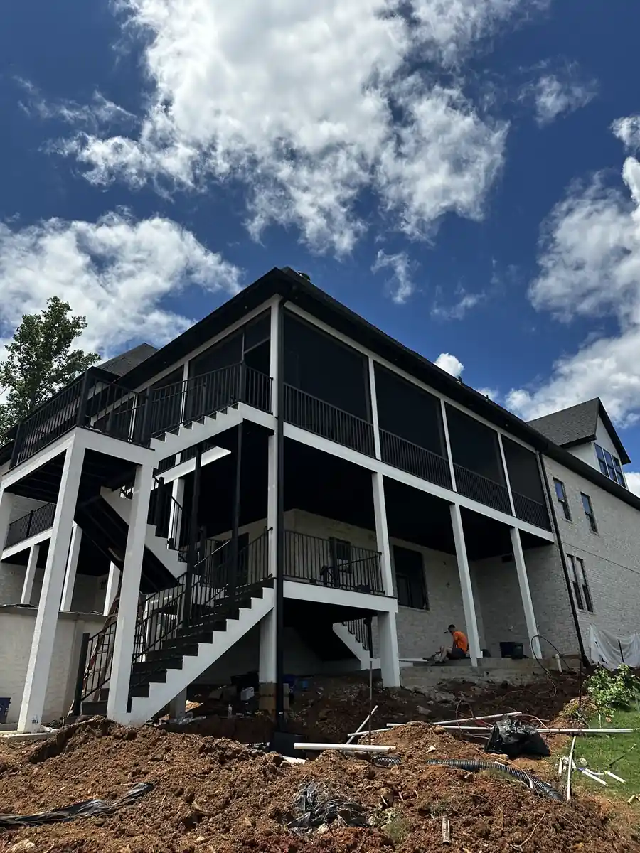 Large light-brick home with an elevated screened upper porch, black rails, and exterior stairs under a blue sky