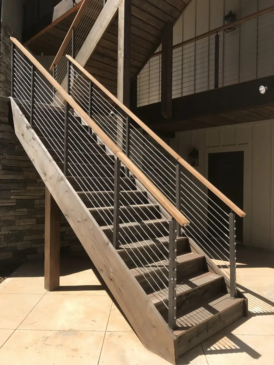 Cable railing with a wood top rail on wood stairs beside a stone wall and concrete patio